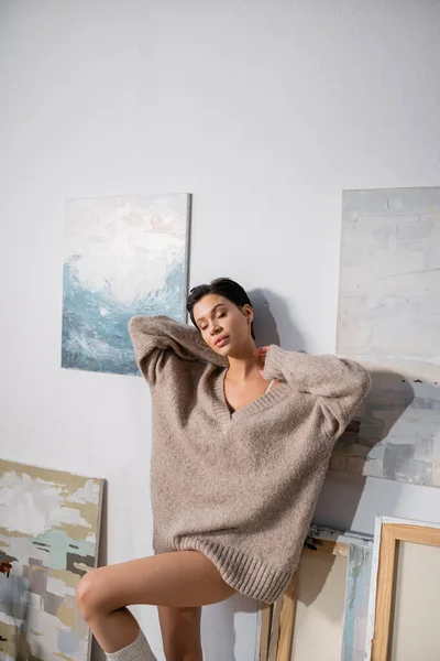 Sensual artist in sweater standing near drawings on wall — Stock Photo