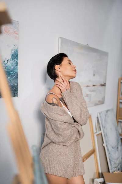 Side view of sexy artist in sweater touching neck near paintings in workshop — Stock Photo