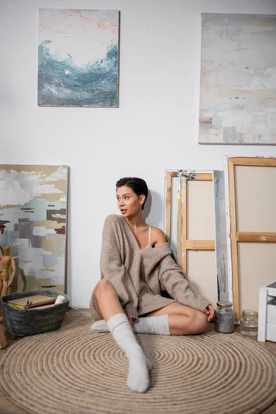 Short haired artist in sweater sitting near paintings on floor in workshop — Stockfoto