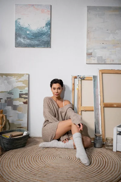 Brunette artist in sweater looking at camera while sitting on floor in workshop — Stockfoto