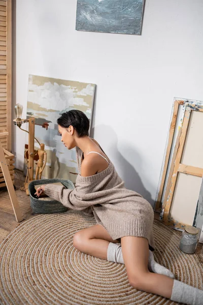 Side view of sexy artist taking paintbrush from basket near paintings in studio — Stockfoto