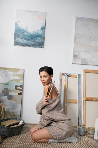 Seductive brunette artist holding paintbrushes while sitting on floor in workshop — Stockfoto