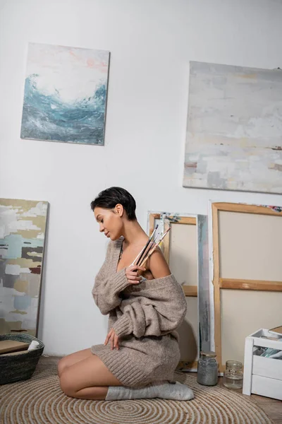 Side view of brunette artist in sweater and bra holding paintbrushes near drawings in workshop — Stock Photo
