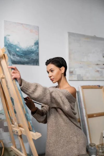Sexy artist in sweater painting on canvas in workshop — Stock Photo