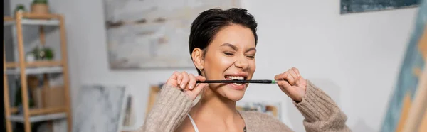 Brunette artist biting paintbrush near blurred canvas in workshop, banner — Stock Photo