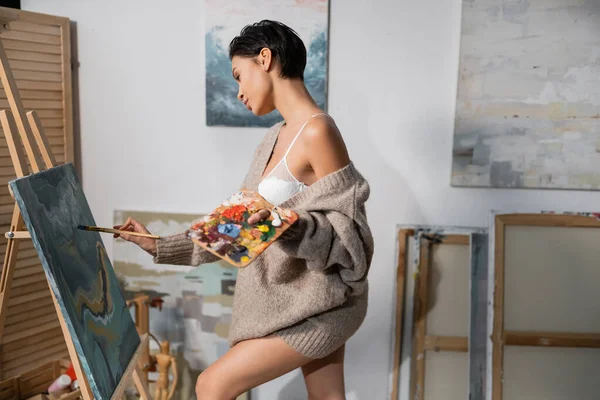 Side view of seductive artist in sweater painting on canvas and easel in studio — Stock Photo