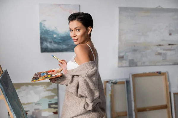 Brunette artist in bra and sweater holding palette with paints near canvas in workshop — Foto stock