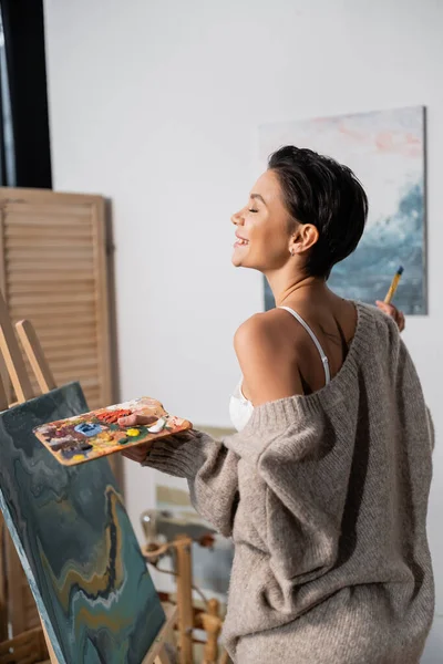 Side view of cheerful artist in bra and sweater holding palette near painting in workshop - foto de stock