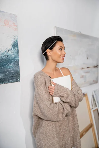 Smiling and sexy artist in sweater with pencil and sketchbook standing in workshop — Stockfoto