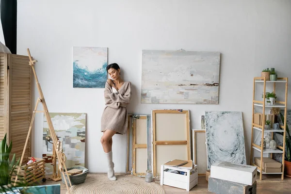 Sexy artist in sweater and socks standing near easel and drawings in workshop - foto de stock