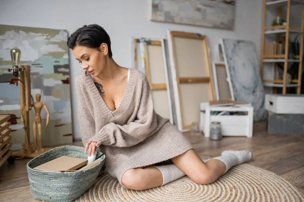 Sexy artist in sweater looking at sketchbooks in basket in workshop - foto de stock
