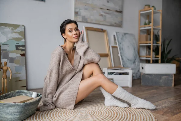 Seductive artist in sweater looking at camera while sitting near basket and paintings in workshop - foto de stock
