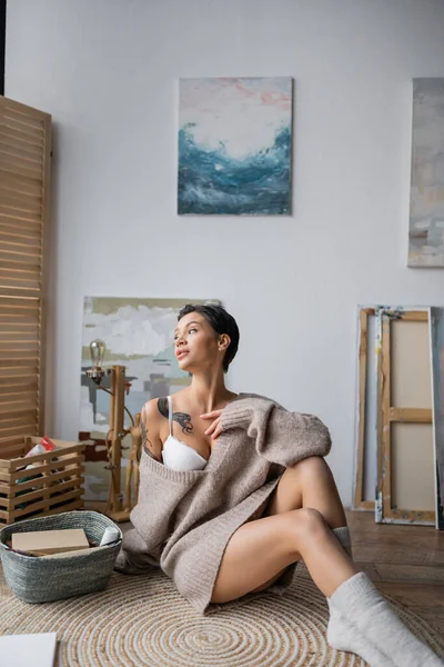 Sexy tattooed artist in bra and sweater sitting near drawings on floor in workshop — Stockfoto