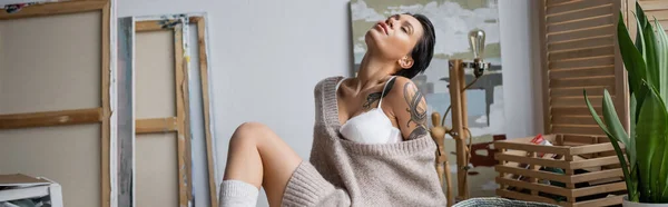 Sexy tattooed artist in bra and sweater sitting in workshop, banner - foto de stock