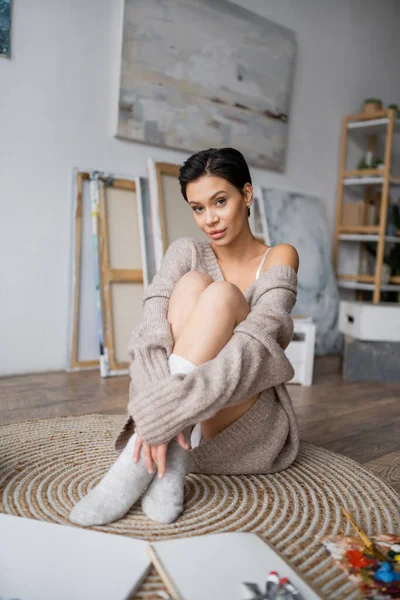 Sensual brunette artist in sweater and socks sitting near sketchbook and palette in workshop — Stockfoto