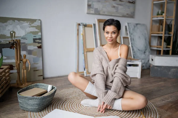 Sensual woman in sweater looking at camera near sketchbook and basket on floor in workshop — Stock Photo