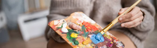 Cropped view of artist in sweater holding paintbrush and palette in workshop, banner — Stock Photo