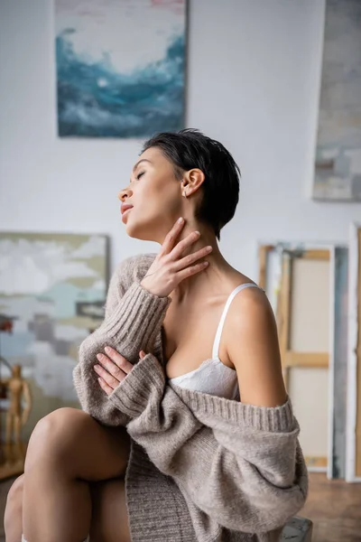 Seductive artist in sweater and bra touching neck in workshop — Stock Photo