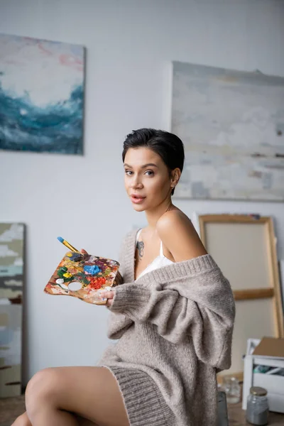 Sensual artist in sweater holding palette and paintbrush in workshop — Stock Photo