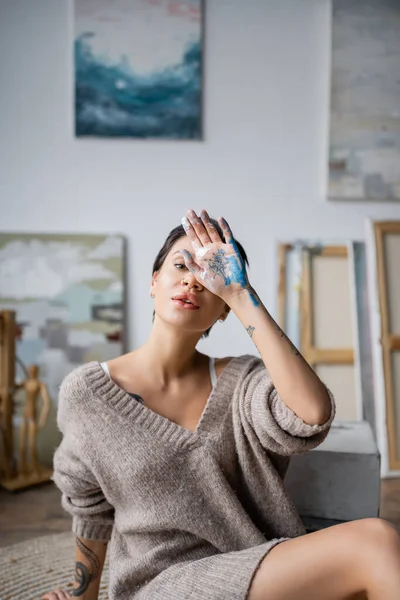 Tattooed artist in sweater holding hand in paint near face in workshop — Stock Photo