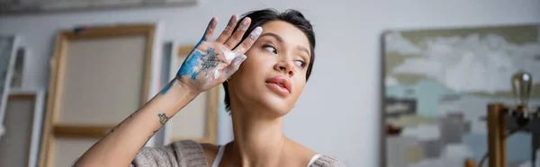 Sexy short haired artist holding hand in paint near face in workshop, banner — Stock Photo