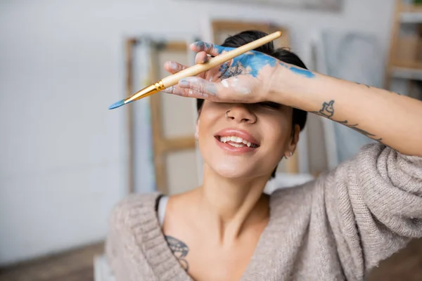 Tattooed artist with paint on hand holding paintbrush in workshop — Stock Photo