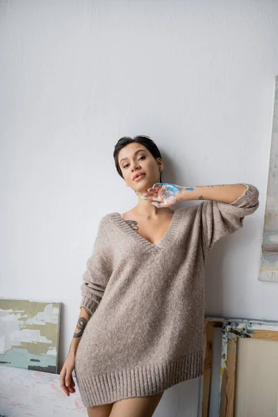 Brunette artist in sweater and paint on hand looking at camera in workshop — Stock Photo