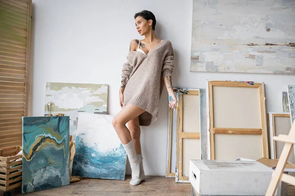Sensual artist in sweater and paint on hand standing near paintings in workshop — Stock Photo