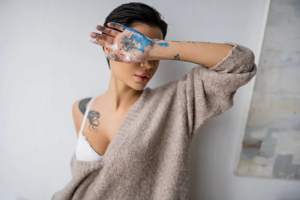 Sensual artist in bra and sweater holding hand in paint near face in workshop - foto de stock