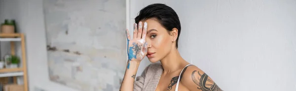 Seductive artist holding hand in paint near face and looking at camera in workshop, banner - foto de stock