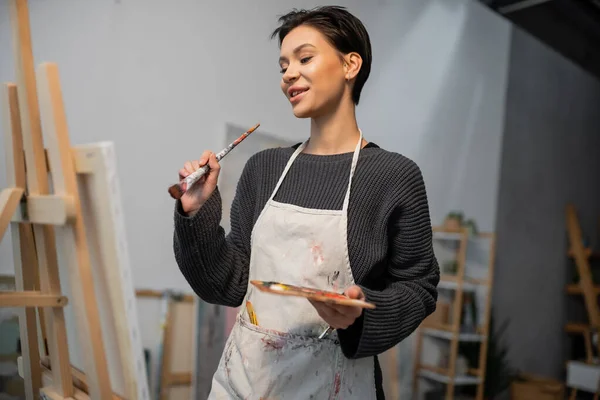 Pleased artist holding palette and paintbrush while looking at canvas in workshop — Stock Photo