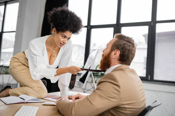 Seductive african american woman sitting on desk and pulling tie of bearded coworker in office — Foto stock