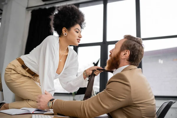 Side view of passionate african american woman sitting on desk and pulling tie of bearded coworker in office — Stock Photo