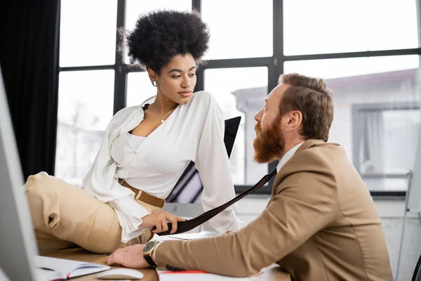 African american woman sitting on desk and pulling tie of coworker in office — Stock Photo