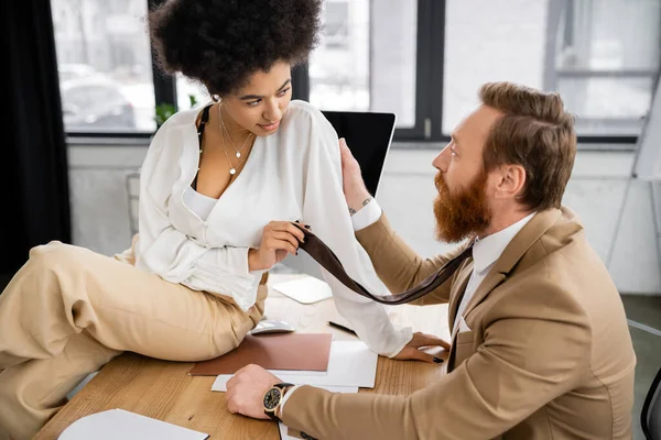 African american woman sitting on desk and seducing tattooed coworker in office — Stock Photo