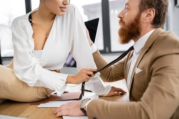 African american businesswoman sitting on desk and pulling tie of tattooed man in office — Stock Photo