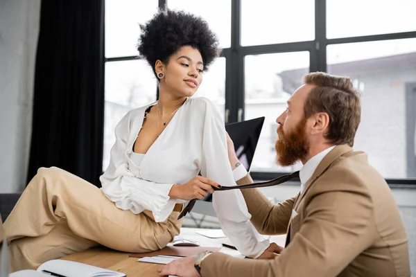 Cheerful african american woman sitting on desk and pulling tie of bearded coworker in office — Stock Photo