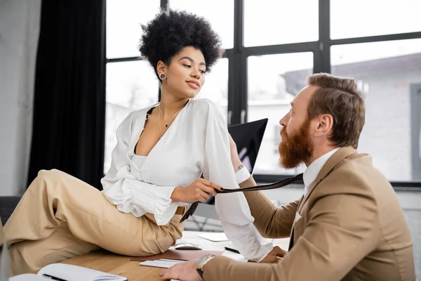 Cheerful african american woman sitting on desk and pulling tie of bearded coworker in office - foto de stock