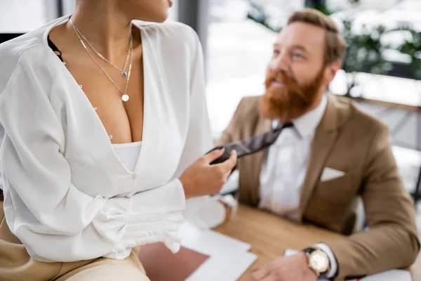 Cropped view of african american woman with big breast sitting on desk and pulling tie of bearded coworker in office — Stock Photo
