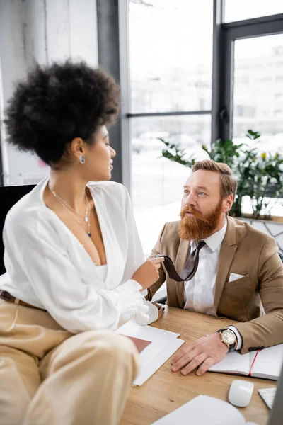 African american woman sitting on desk and pulling tie of bearded coworker in office — Stock Photo