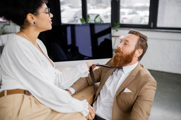 Cheerful african american woman in glasses pulling tie while seducing bearded coworker in office — Fotografia de Stock