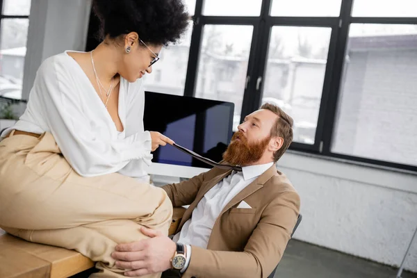 Smiling african american woman in glasses pulling tie while seducing bearded coworker in office — Photo de stock
