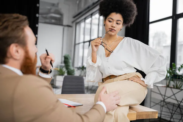 Sexy african american businesswoman holding glasses and posing while sitting on desk near bearded coworker — Stock Photo