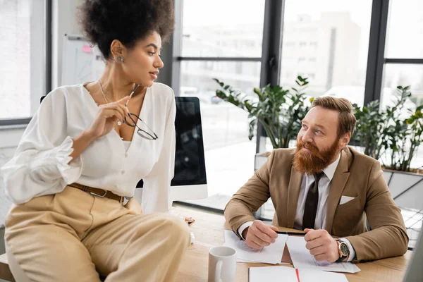 Seductive african american businesswoman holding glasses and sitting on desk near smiling businessman — Stock Photo