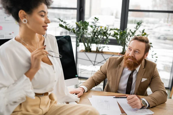 Cheerful and bearded man looking at seductive african american businesswoman holding glasses while sitting on desk — Stock Photo