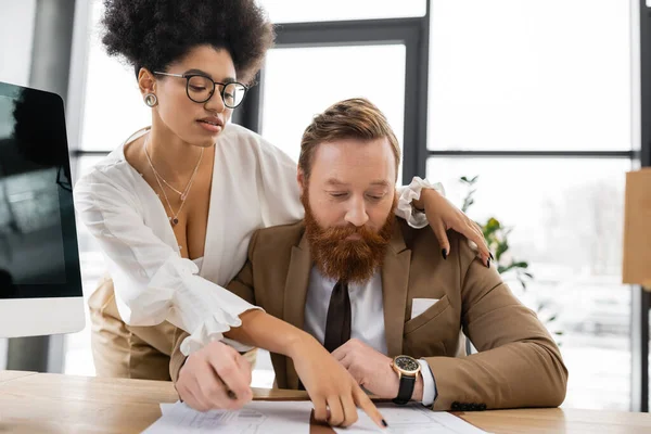 Seductive african american businesswoman hugging bearded coworker and pointing at document on desk — Stock Photo