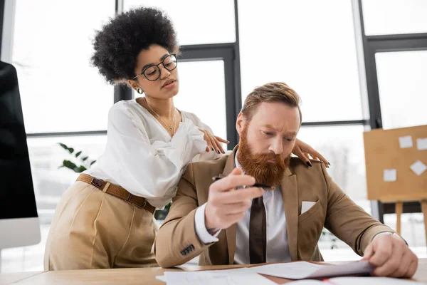 Curly african american businesswoman touching shoulders of businessman working with documents in office — Stock Photo