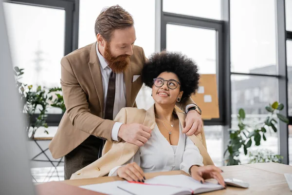 Bearded businessman wearing jacket on cheerful african american woman in office — Stock Photo