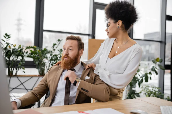 Seductive african american woman touching jacket of surprised man in office — Stock Photo