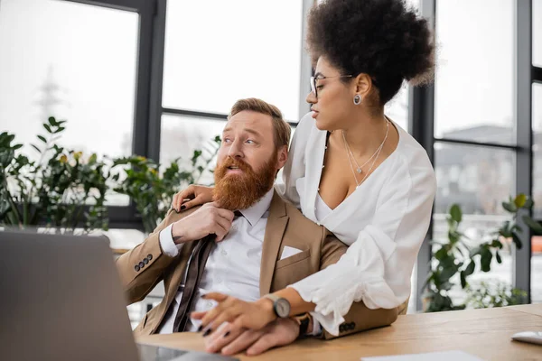 Seductive african american woman using laptop near bearded businessman pulling tie in office — Stock Photo